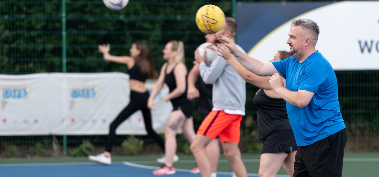 A man throws a netball during practice on an outdoor court, with a row of other adults doing the same in the background.