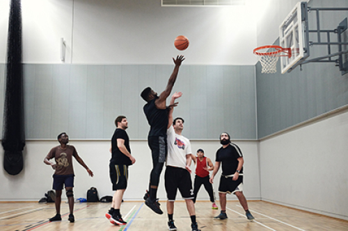 A man shoots a basketball over another player who tries to block it, while other players watch on