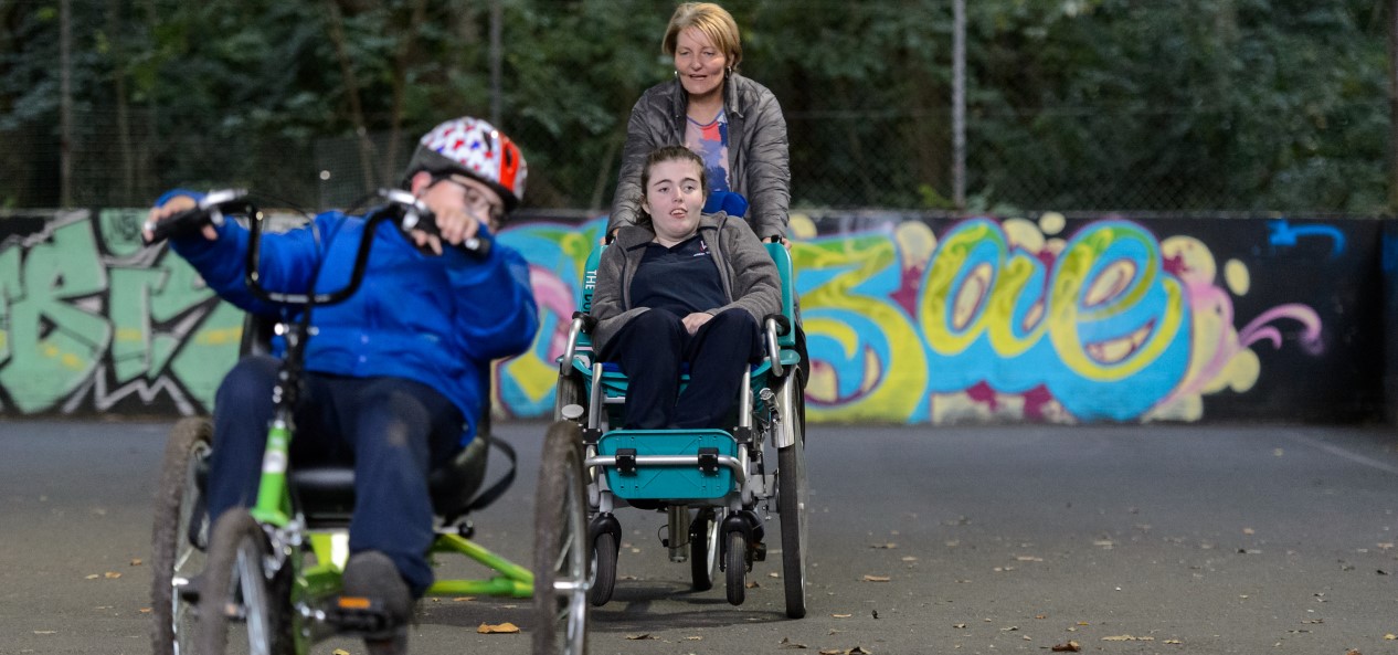 A boy rides an adaptive three-wheel bike ahead of a girl in a similar bike being pushed by a woman.