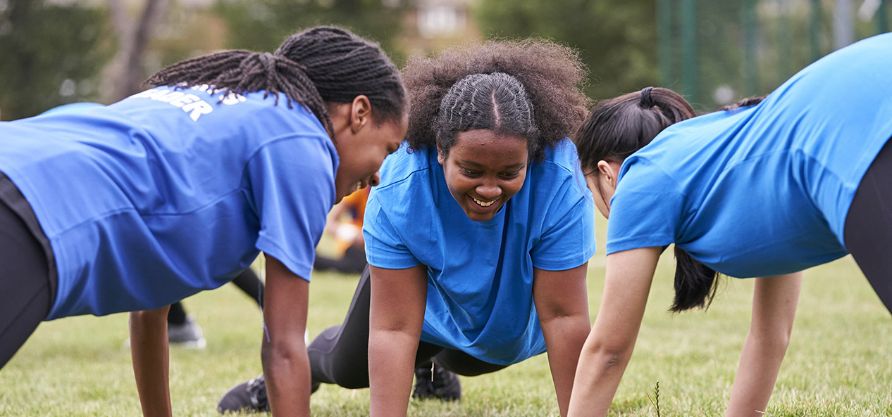 Three young girls adopt a press-up position on the grass outside, all facing each other in a circle