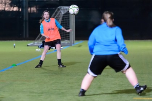 Two young female footballers with the ball in the air on an outdoor artificial grass pitch at night.