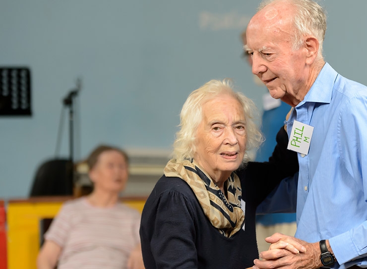 An elderly man and woman dance, arm in arm, in a community centre