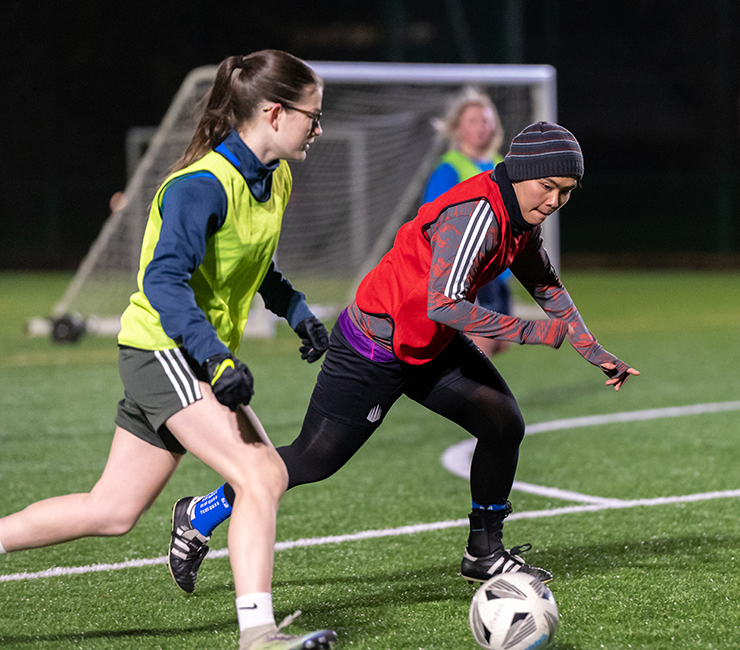 Women wearing training bibs of different colours play football during an evening training session outdoors.