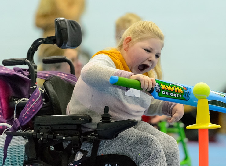 A girl in a wheelchair plays tee-cricket in a sports hall