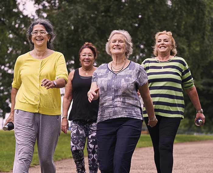 Four older ladies in active outfits smile as they walk next to each other in a park.