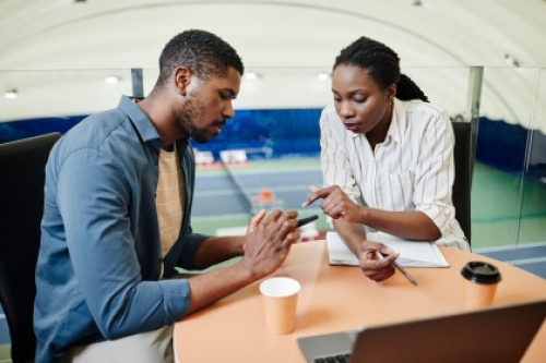 Two people consider something on a mobile phone while sat at a work desk above indoor tennis courts.
