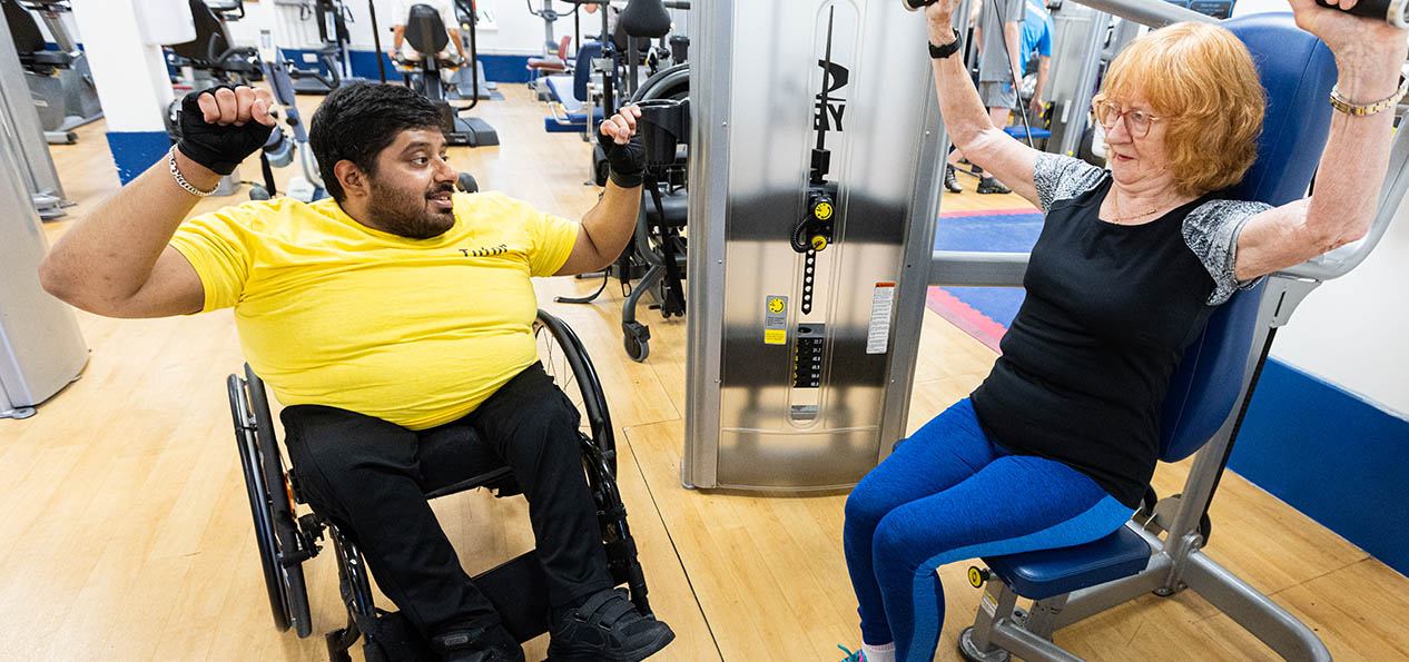 A young man on a wheelchair coaches an older lady during a machine session in a gym.
