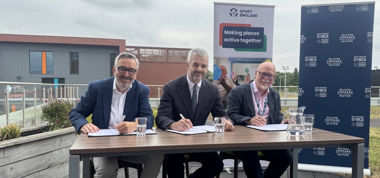 Sport England chief executive Tim Hollingsworth, South Yorkshire mayor Oliver Coppard, and South Yorkshire Integrated Care Board CEO Gavin Boyle sign a memorandum of understanding at a table outside.