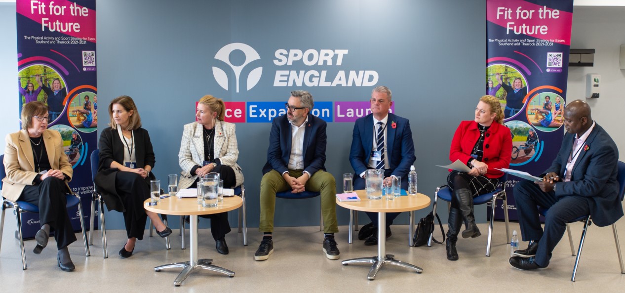 Tim Hollingsworth sits in a row of seven people at Sport England's Place Expansion launch, with banners in the background.