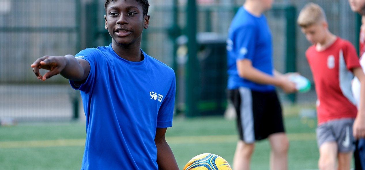 A Black boy wearing a blue t-shirt with a white Youth Sport Trust logo stands on an outdoors football pitch pointing at something with his right hand while holding a yellow football with the other one. A group of three kids is seen behind him.