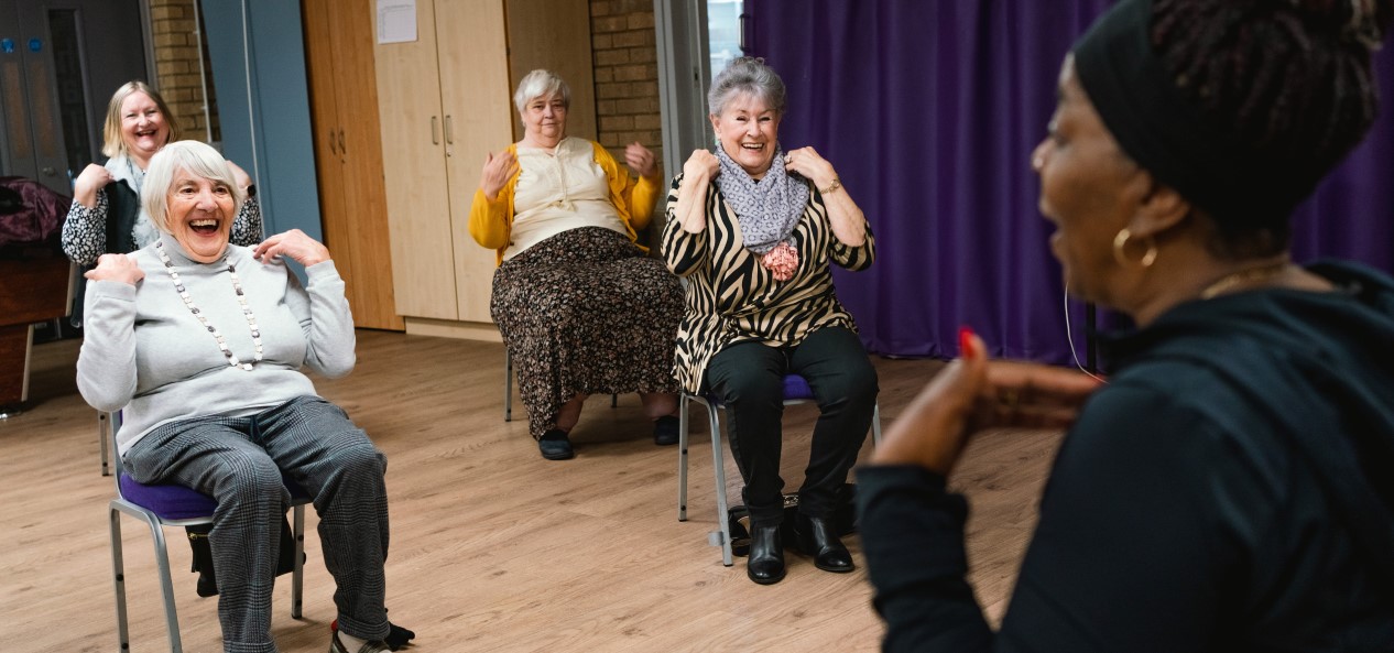 Four older women touch their shoulders while taking part in a seated exercise session with a trainer in a hall.