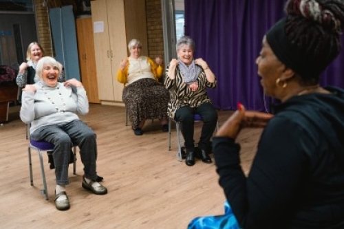 Four older women touch their shoulders while taking part in a seated exercise session with a trainer in a hall.