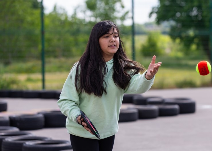 A girl holding a velcro ball-catching pad throws a ball in a playground.