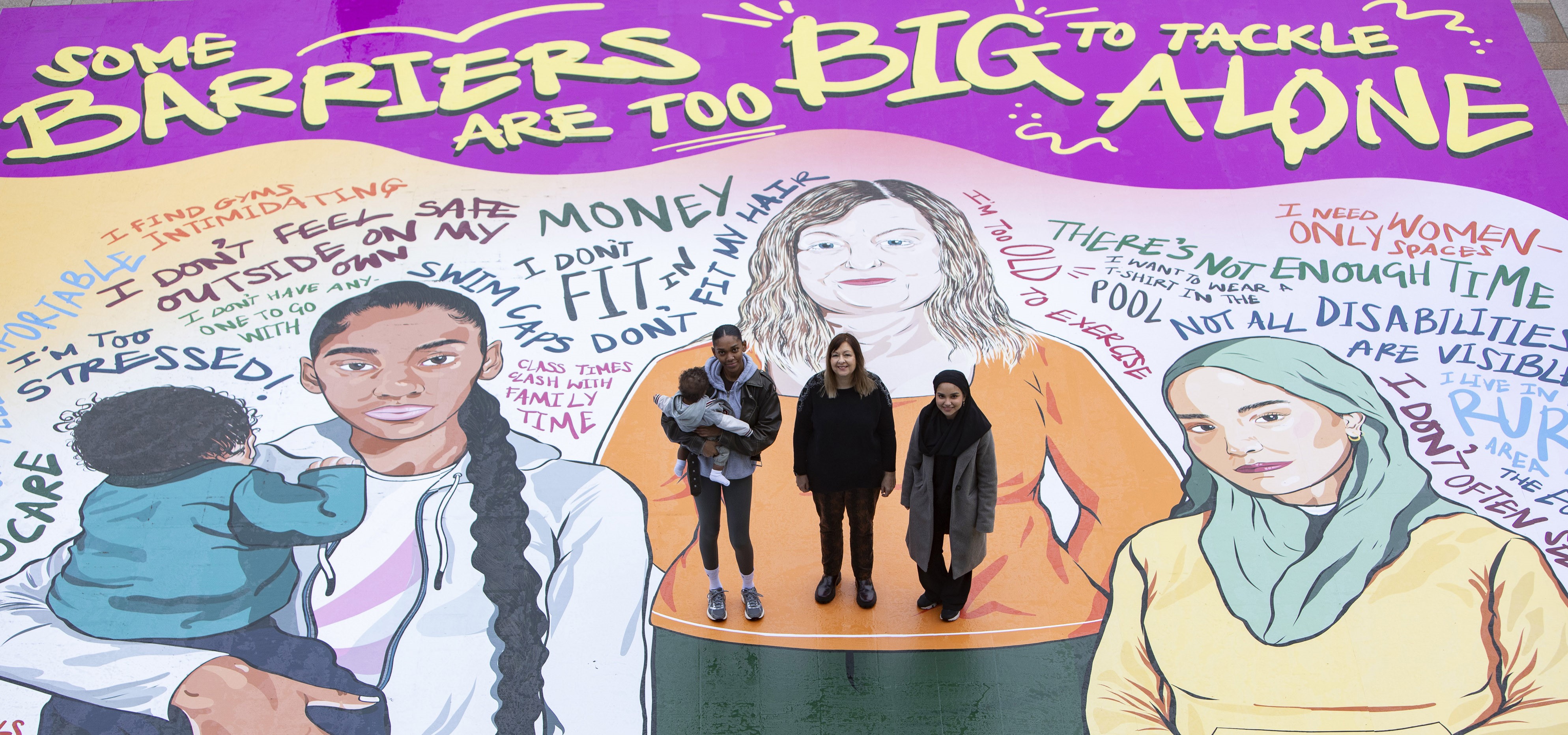 Three women, one holding a baby, stand on a giant mural featuring illustrations of women and the text 'Some barriers are too big to tackle alone'.