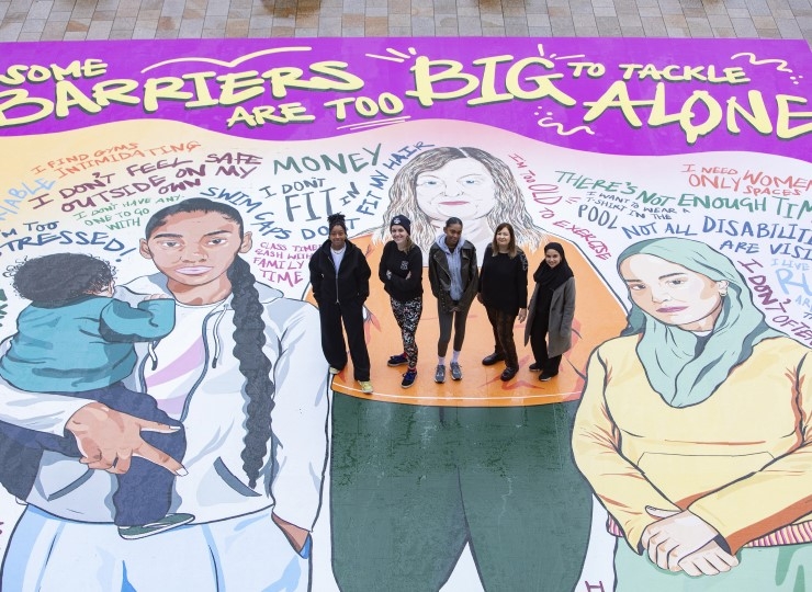 Five women stand on a giant mural featuring illustrations of women and the text 'Some barriers are too big to tackle alone'.