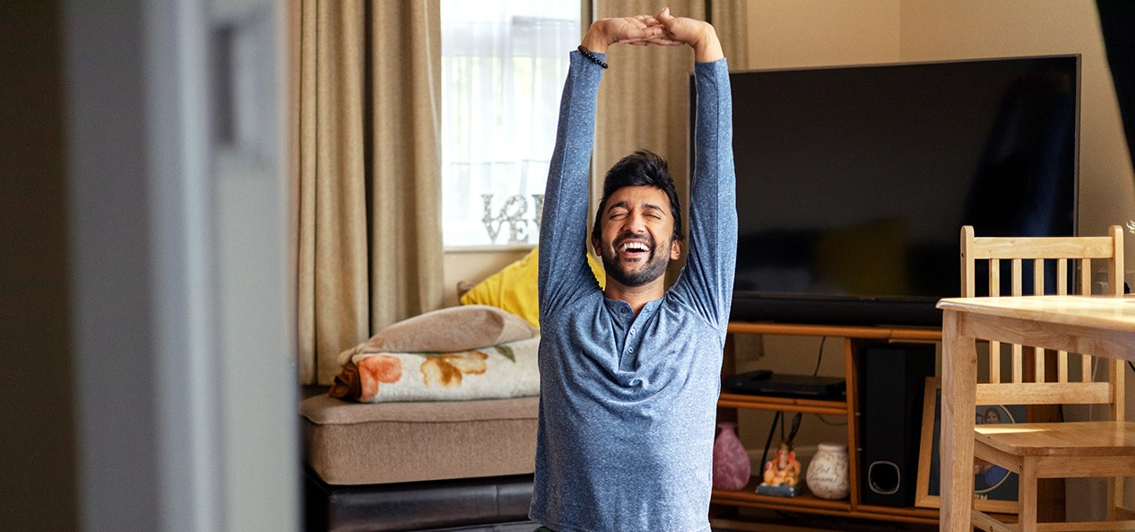 A man smiles with his eyes closed as he stretches upwards during a yoga session in his living room.