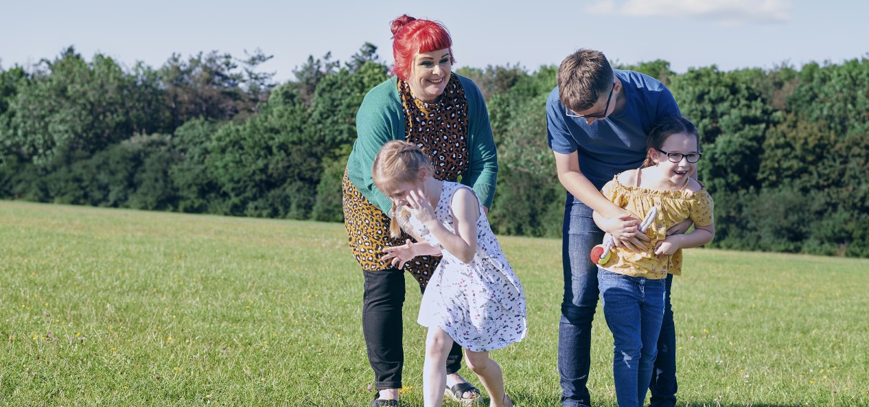 A man and a woman playfully reach out to two laughing girls on a field on a sunny day.