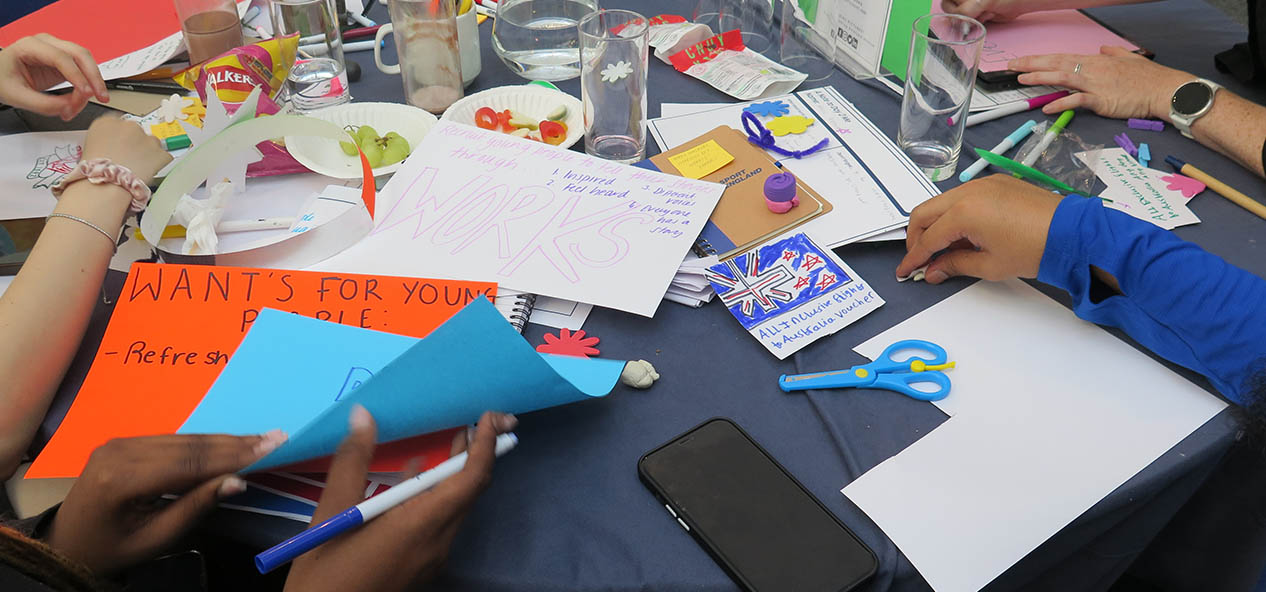 The hands of young people are seen working on some papers on a table where there's also some grapes, a bag of crisps, a pair of scissors, glasses and colour pens. 