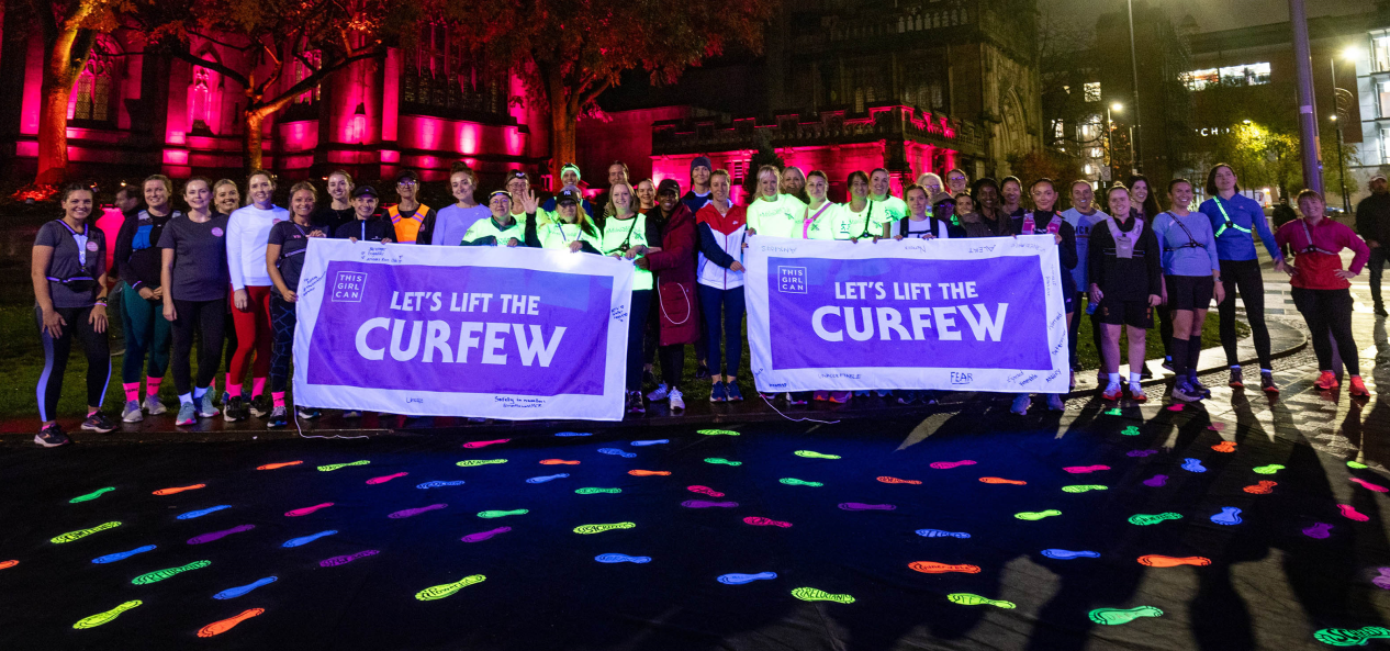 Runners pose with Let's Lift the Curfew signs in front of glow-in-the-dark footprints at night in Manchester